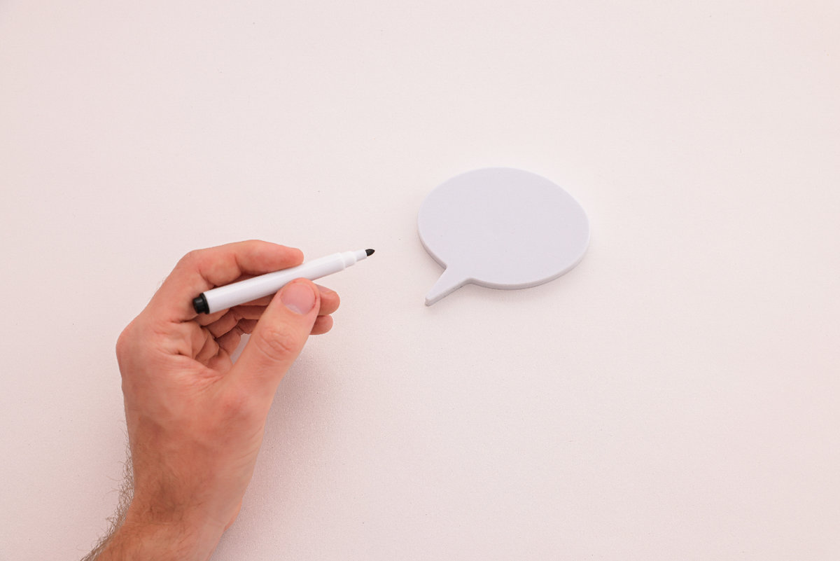 A white speech bubble sits on a white background. A hand holding a dry erase marker hovers above the speech bubble, as if it is going to write on the bubble.